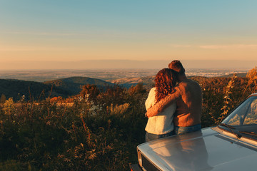 Rear view of beautiful young couple resting after road trip leaning against car and enjoying the...