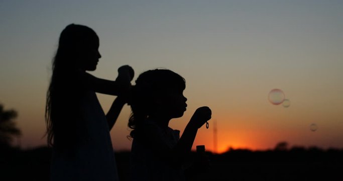 Silhouette of two pretty  girls of four and seven years old with long hair blowing  bubbles in the sunset of a warm summer day against a bright orange sky. 4k slow motion 50 fps