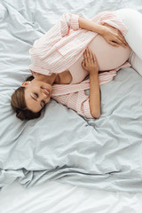 Fototapeta na wymiar overhead view of smiling pregnant woman lying in bed and touching belly