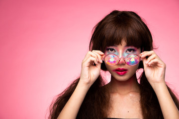 Fashion Model looks at camera for shooting new collection Sunglasses.  Beautiful Asian Woman summer trend make up wear Kaleidoscope Glasses with colorful, studio lighting pink background