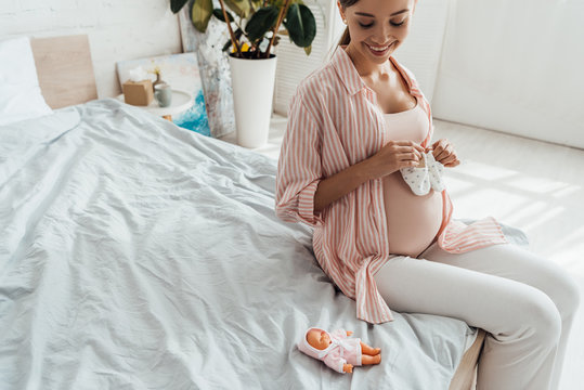 smiling pregnant woman sitting on bed and holding baby shoes