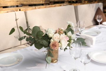 Wedding table setting decorated with fresh flowers in a brass vase. Wedding floristry. Banquet table for guests outdoors with a view of green nature. Bouquet with roses, eustoma and eucalyptus leaves