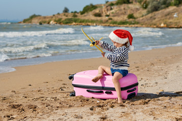 Little girl in Santa hat playing with toy airplane on sunny tropical beach. Child sits on pink suitcase. Christmas and new year vacation travel concept