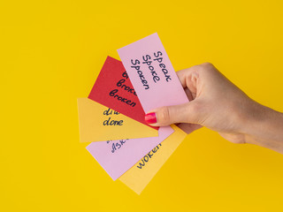 Female hand holds flashcards with handwritten irregular English verbs isolated on yellow. Language learning concept