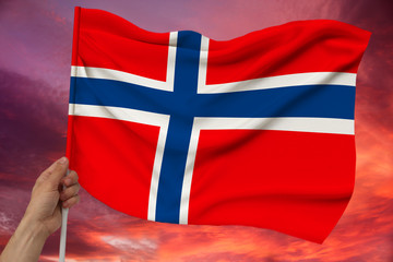 hand holds against the background of the sky with clouds the colored flag of Norway on the texture of the fabric, silk with waves, close-up