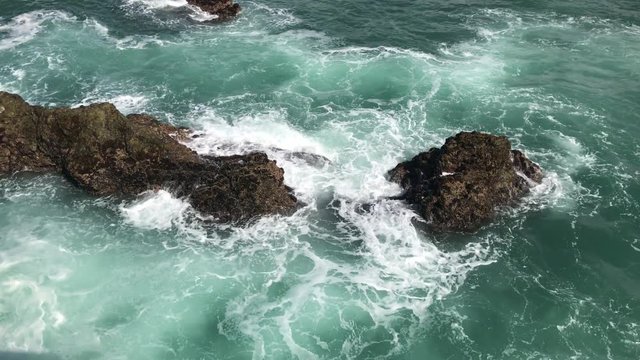 Waves in strong swirling undertow current violently splashing emerald green salt water and sea foam mist on rough coastal rocks on a hot summer day