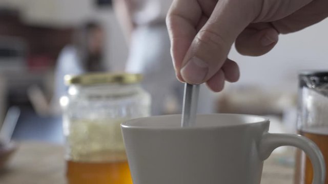 A man starts to stir, mixing a mug of hot tea in the kitchen, blurred people talking in the background.