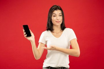Naklejka premium Caucasian young woman's half-length portrait on red studio background. Beautiful female model in white shirt. Concept of human emotions, facial expression, sales. Showing on phone, looks confident.