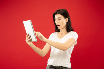 Caucasian young woman's half-length portrait on red studio background. Beautiful female model in white shirt. Concept of human emotions, facial expression. Using tablet for making her vlog or talking.