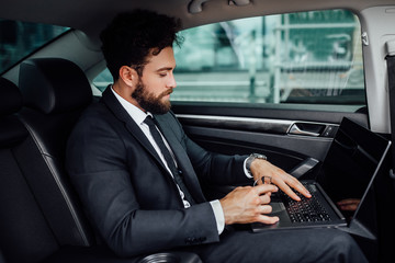 Handsome, bearded, smiling top manager in black suit working on his laptop on the backseat of the car.