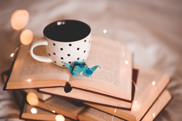 Vintage cup of tea with paper origami crane on open books over lights close up. Night time. Good morning.