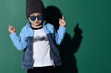 Portrait of cool kid boy in blue sunglasses, hat, fleece jacket and white t-shirt cool dancing on green