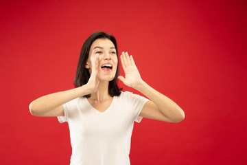 Caucasian young woman's half-length portrait on red studio background. Beautiful female model in white shirt. Concept of human emotions, facial expression. Calling somebody for sales and purchases.