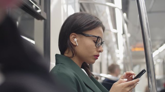 Close up of young businesswoman sitting in subway car, holding telephone in hands, using it and listening to audio book via wireless headphones