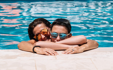 Mother and son with sunglasses are leaning on the edge of the pool on a sunny summer day.