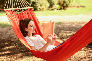 Beautiful young woman with book resting in hammock outdoors