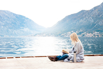 Fototapeta premium Woman is reading book on wooden pier by sea, mountains, beach. Cozy winter picnic with coffee, hot beverages or tea in thermos, warm plaid. Girl is enjoying life, relaxation, wellbeing.