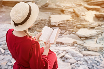 An attractive Caucasian girl in a red bright dress and a straw hat sits on a large stone by the sea at sunset and reads an interesting book on the sea horizon. Back view