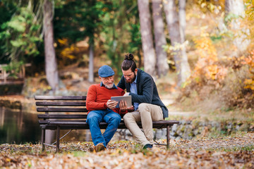 Senior father and his son sitting on bench in nature, using tablet.
