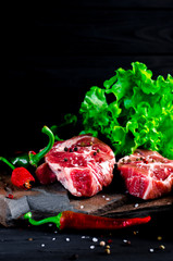 Two juicy steaks lie on a wild stone next to red and green chili peppers and a fresh green salad on a black wooden table
