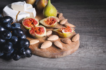 Still life cheese plate on a wooden cutting board lies sliced cheese with mold almonds in the shell and sliced figs next to a bunch of black grapes. Delicious and healthy food concept
