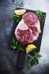 Black wooden chopping board with raw cross cut veal shank for cooking ossobuco, flatlay on a grey stone background