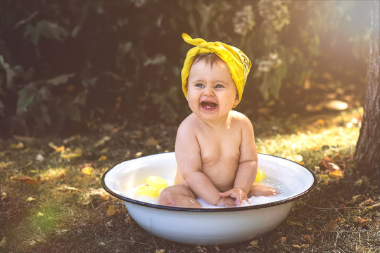 Baby kleines M&auml;dchen Kind sitzt in Emaillle Badewanne im Garten mit gelbem Stirnband lacht Sommer