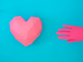 A pink paper heart of multiple faces, crafted in low poly technique, combined to a pink gloved hand, over a contrasting background