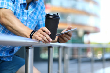 Focus on male hands holding cup of coffee and tablet. Guy standing leaning on railing and surfing internet via gadget. Modern technology concept. Blurred background