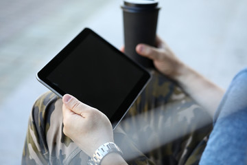 Focus on male hands holding stylish gadget and paper cup of coffee in hands. Guy surfing internet, chatting, messaging via special app on notepad. Copy space on empty screen. Blurred background
