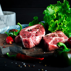 Two juicy steaks lie on a wild stone next to red and green chili peppers and a fresh green salad on a black wooden table