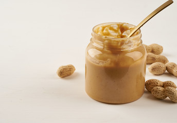 jar of peanut butter and peanuts in shell on white table, side view, fresh ground crushed nuts,...