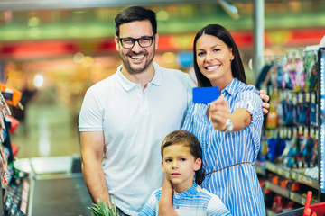 Obraz premium Happy family with child and shopping cart buying food at grocery store or supermarket, woman holding credit card.