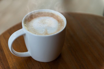 Close up White cappuccino coffee cup With art foam on a brown wood table near a light-colored window on the table at the coffee shop.
