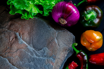 A wild multi-colored stone with a place for text lies on a black table next to fresh blue onions, yellow peppers, chili and lettuce