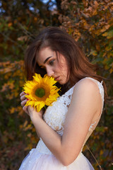 Beautiful lovely girl in a white dress sniffs a flower of a sunflower. Sunlight is playing. Enjoying nature. Autumn time.