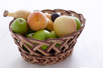 Apples, pears, plums, banana and peach in a wicker, brown basket on a light background. Fruit on a white background. Background of fruits.