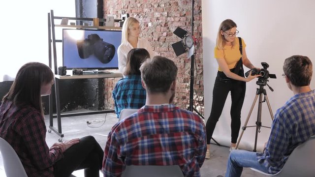 photographer talks about device of a DSLR camera to young creative people at a photo seminar, in a professional studio on background of TV with picture of equipment