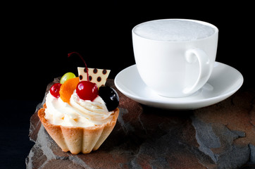 Fruit cake with cherries, grapes and plum stands on a wild stone next to a white cup of coffee with a foam