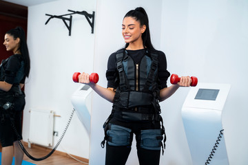 Pretty female athlete exercising with small dumb bells on ems machine 
