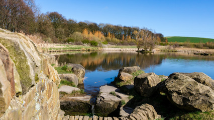 Fototapeta na wymiar Herrington Country Park in Sunderland. Image features trees, rock wall and a lake that is reflecting a cloudless sky on a spring/summer day.