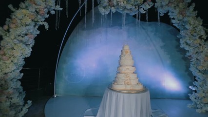 Beautiful tasty wedding cake under decorated arch against blue background at night. Steadicam shot