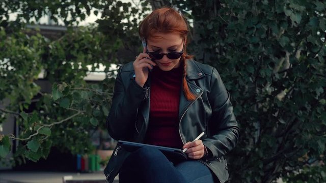 Young woman in sunglasses talking on the mobile phone while sitting in the Park on a bench, records of conversation in notes on a tablet. Girl talking on the phone.