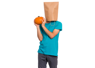 Happy halloween concept. Teen boy with paper bag over his head holds orange small fresh pumpkin, isolated on white background. Teenager cover head with bag. Child with food - raw vegetables in hands.