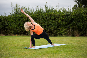 A middle aged woman practicing yoga barefoot outside in a grassy park. She is wearing a bright orange vest and multi coloured leggings. The style of yoga she is doing is  Hatha Yoga