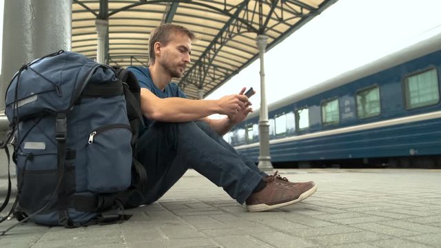 Young bearded man tourist with smartphone and backpack sitting on railway station platform and waiting for train. Travel concept.