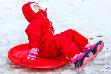 Pretty smiling little girl in her ski suit sliding down a small snow covered hill with her sledge