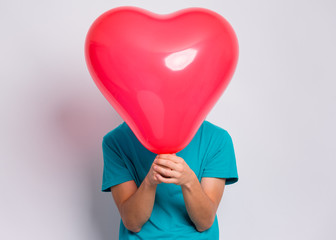 Teen boy hiding behind red heart shaped balloon. Child holding symbol of love, family, hope. Celebration of Saint Valentines Day. Teenager cover face posing in studio.
