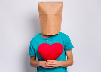 Valentines Day concept. Teen boy with paper bag over head holds red heart. Boy holding symbol of love, family, hope. Teenager cover head with bag posing in studio. Child pulling paper bag over head.