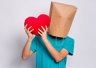 Valentines Day concept. Teen boy with paper bag over head holds red heart. Boy holding symbol of love, family, hope. Teenager cover head with bag posing in studio. Child pulling paper bag over head.